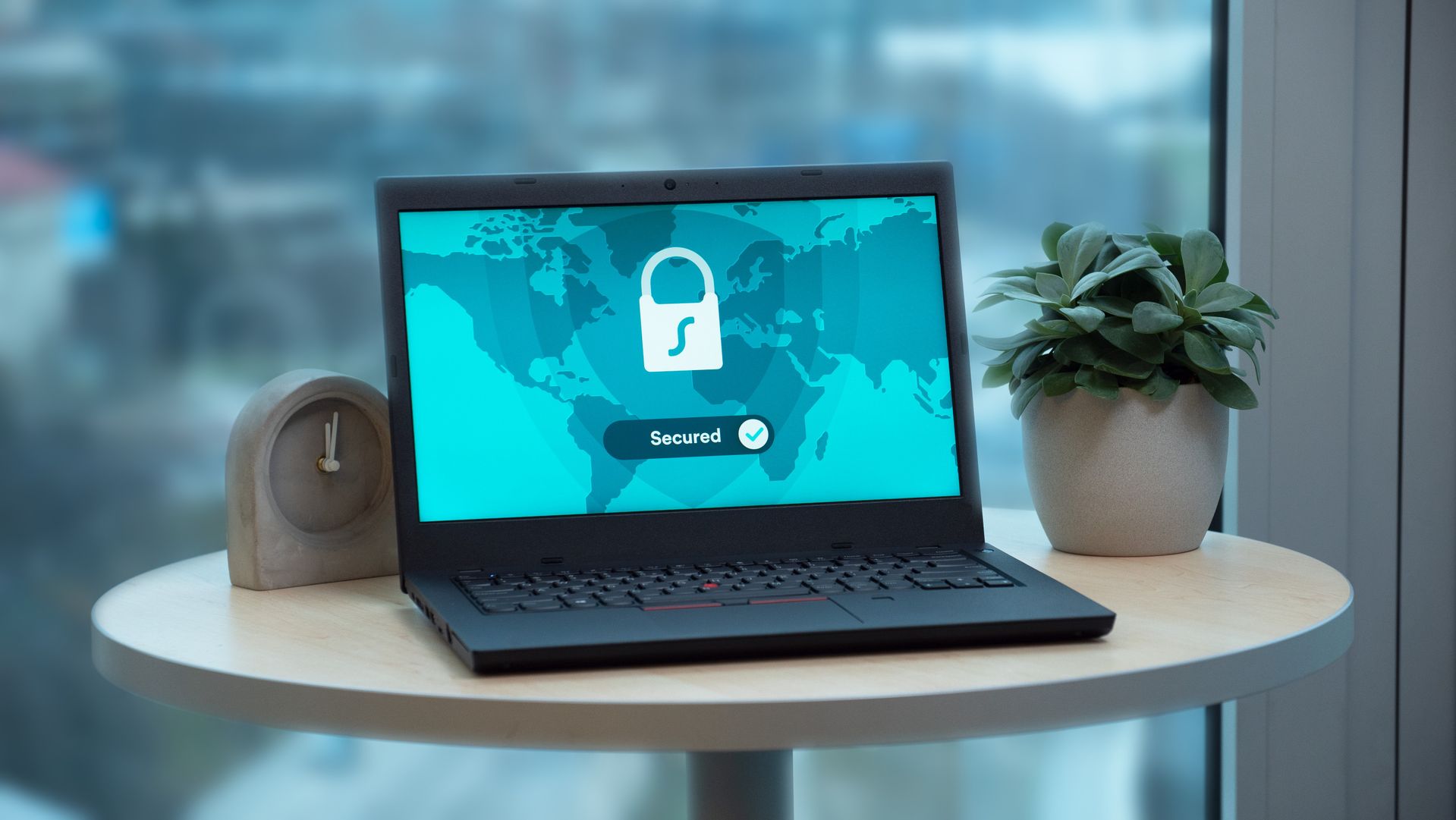 Laptop on table showing lock icon and Secured text over world map, beside a small potted plant and clock near a window