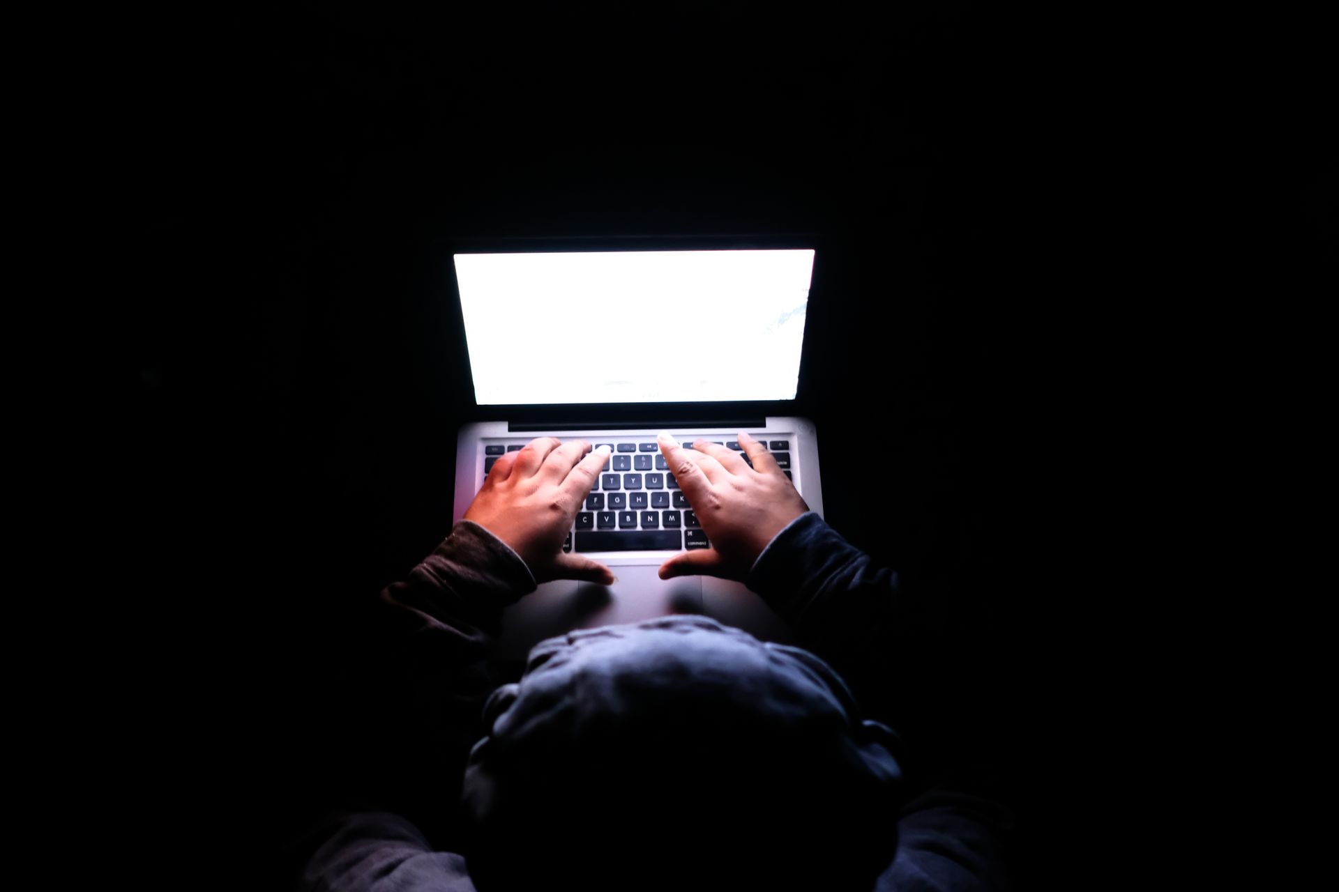 Top-down view of an anonymous person in a dark room typing on an illuminated laptop with a bright, glowing white screen